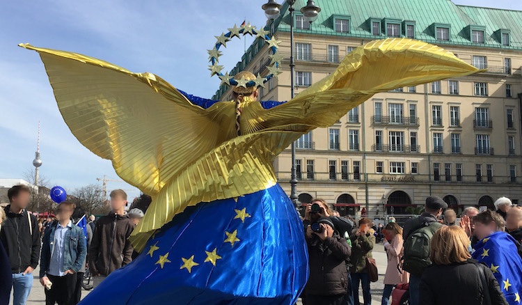 Tanzende Figur mit Europa-Kostüm mit Goldflügeln. Im Hintergrund ist der Berliner Fernsehturm zu sehen.