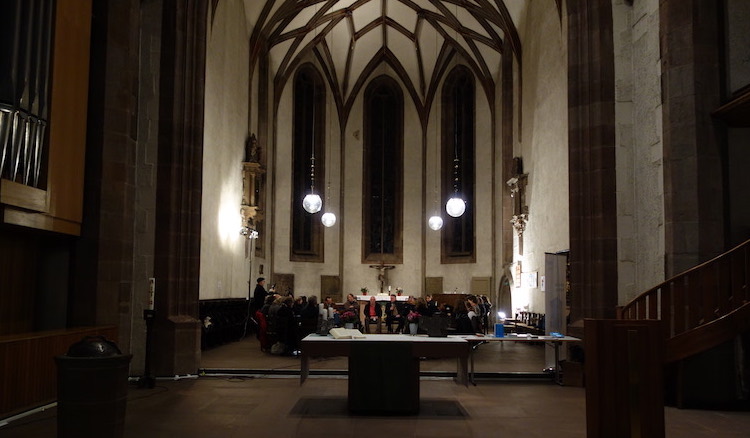 Blick in den großen recht dunklen Kirchenraum in dem hinten unter vier Leuchtern die Runde von ca. 30 Leuten im Kreis sitzt und diskutiert