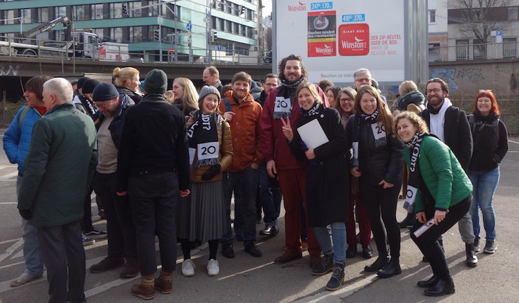Circa 30 Leute stehen inmitten des tiefergelegten Parkplatzrondells unten am Österreichischen Platz und Grinsen um die Wette